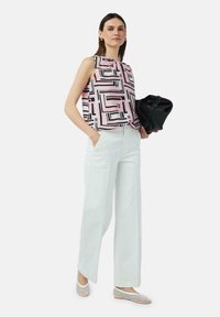 Mouwloze blouse in het roze met zwart geometrisch patroon, gecombineerd met lichtgroene wijde broek en metallic platte schoenen; houdt een zwarte tas vast.