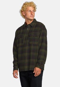 Camisa de franela verde con patrón de cuadros rojos y amarillos, dos bolsillos en el pecho, cierre de botones en la parte delantera y mangas largas. Fabricada en un tejido suave y texturizado.