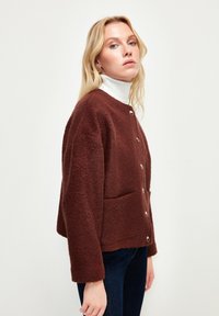 Brauner Fleece-Jacke mit rundem Ausschnitt, Fronttaschen und Akzenten mit Silberknöpfen. Getragen über einem weißen Rollkragenpullover.