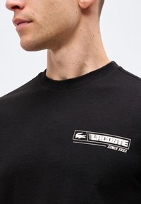 Νέος άντρας με κοντά μαλλιά φοράει μαύρο T-shirt Lacoste με λευκό λογότυπο και κείμενο "Since 1933" στο στήθος.