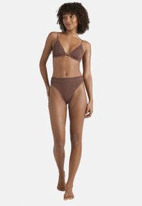 Bikini marrone lavorato a maglia con top triangolare e slip a vita alta. Presenta spalline sottili e un motivo testurizzato. La modella è in piedi a piedi nudi.