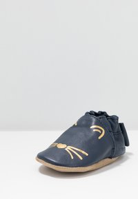 Pantofle pentru bebeluși din piele bleumarin, cu o talpă moale din suede, având accente aurii în formă de față de pisică, inclusiv mustăți și un nas în formă de inimă.
