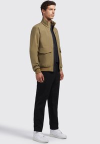 Khaki jas met een hoge kraag; twee voorzakken; gecombineerd met donkere broek en witte sneakers; soepel stof met een moderne pasvorm.