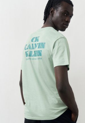 Homme à la peau foncée et aux dreadlocks tournant la tête par-dessus l'épaule, portant un t-shirt Calvin Klein vert clair avec un logo bleu dans le dos.