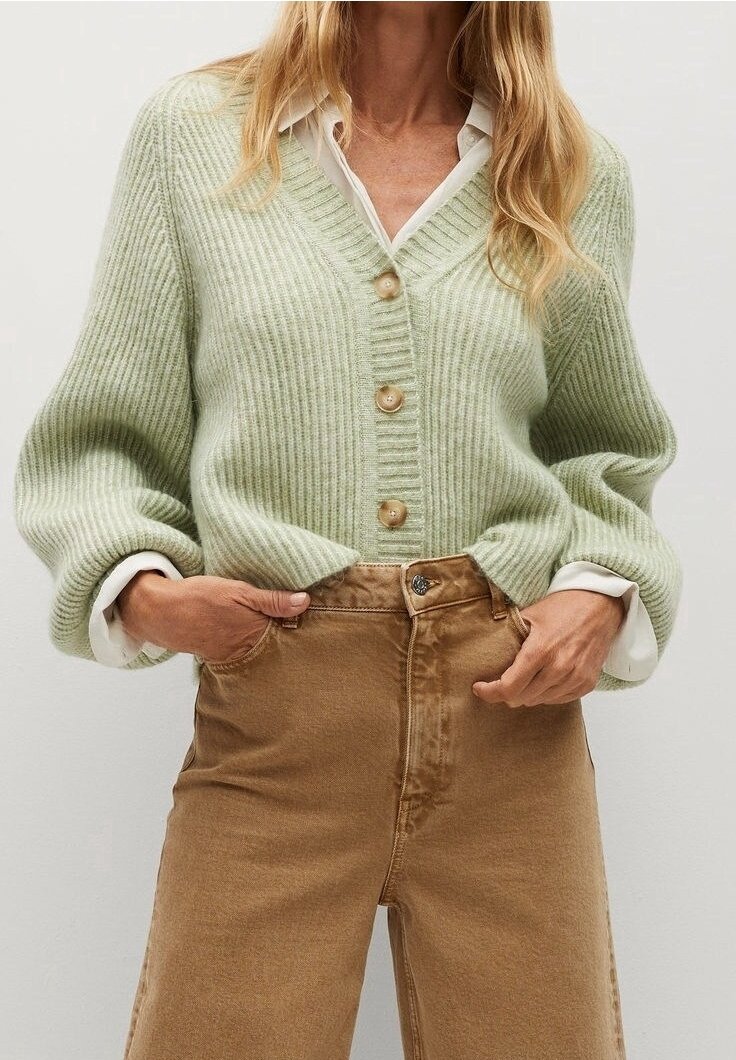 Femme portant un cardigan côtelé vert clair à boutons en bois sur une chemise blanche, associé à un pantalon marron taille haute, les mains dans les poches.