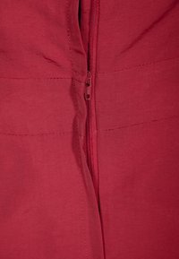 Roter Stoff mit texturierter Oberfläche, ausgestattet mit einem Nylonreißverschluss in der Mitte für den Verschluss. Nähte sind entlang der Nähte sichtbar.