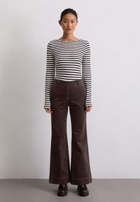 Vrouw die tegen een effen muur staat, gekleed in een zwart-wit gestreepte longsleeve top, donkerbruine wijde broek en zwarte schoenen.