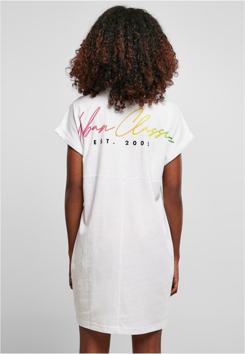 Femme aux cheveux bouclés portant une robe blanche à manches courtes avec l'inscription « Urban Classic EST. 2005 » en couleurs arc-en-ciel dans le dos.