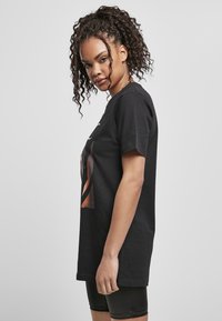 T-shirt en coton noir avec un imprimé graphique sur le devant. Le modèle se tient de profil, mettant en valeur la coupe décontractée et les manches courtes.