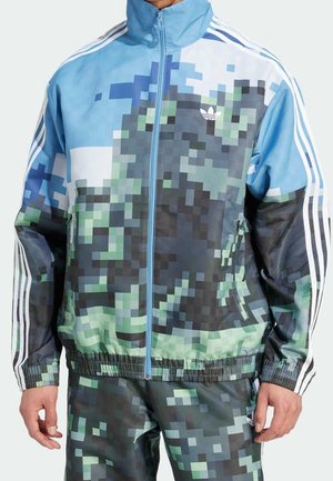 Uomo che indossa giacca e pantaloni Adidas a camouflage pixelato blu e verde con dettagli a tre strisce bianche sulle maniche e sulle gambe.