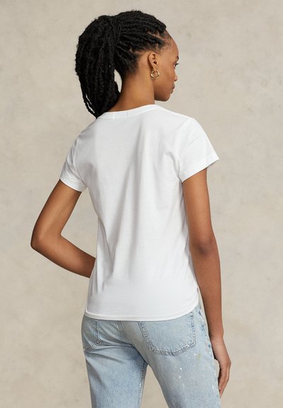 Kvinna med dreadlocks som bär guldörhängen, en enfärgad vit t-shirt med kort ärm och ljusblå jeans med färgsplatter, vänd bort.