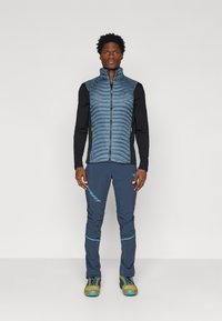 Αυθεντικό ελαφρύ αδιάβροχο γιλέκο σε σκούρο μπλε με κατακόρυφες ραφές, συνδυασμένο με σκούρες μπλε παντελόνες που διαθέτουν μπλε λογότυπο και λεπτομέρειες. Υπαίθρια υποδήματα σε πράσινο χρώμα.