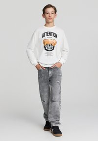 Biała koszula z długim rękawem z grafiką przedstawiającą misia i czaszkę, w połączeniu z zniszczonymi szarymi dżinsami i czarnymi sneakersami.