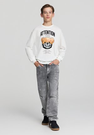 Camicia bianca a maniche lunghe con una grafica di un orso di peluche e un teschio, abbinata a jeans grigi strappati e sneakers nere.