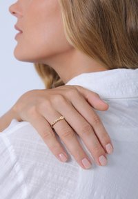 Goldring mit mehrfarbigen Steinen, der ein schlankes Design aufweist. Die Hand liegt auf einem weißen Textilhemd, das natürlichen Nagellack zeigt.