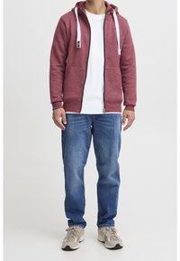 Rode hoodie met witte koorden, ritssluiting en voorzakken. Gecombineerd met blauwe jeans en beige sneakers. Eenvoudig, casual ontwerp.