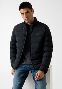 Schwarze gesteppte Jacke mit hohem Kragen, Reißverschluss und Logodetails. Kombiniert mit einem grauen Shirt und blauen Jeans. Glatter Stoff.