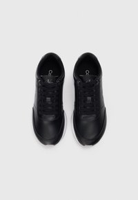 Un paio di sneakers nere Calvin Klein con lacci, suole bianche e logo del marchio visibile sulla linguetta, viste dall'alto su uno sfondo grigio.