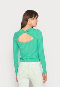 Camisola de manga longa verde com recorte nas costas. O tecido parece macio; o corte justo destaca a silhueta. Combinada com jeans verde claro.