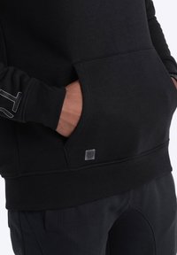 Černá mikina s kapucí vyrobená z měkké tkaniny, vybavená klokaní kapsou a logem. Elastické manžety a lem dotvářejí moderní design.