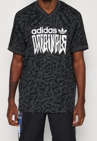 Černé tričko s krátkým rukávem a geometrickým vzorem, s výrazným bílým logem "adidas ORIGINALS" na přední straně a designem ve tvaru V.