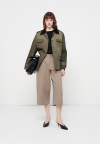 Jachetă verde măsliniu cu guler negru, două buzunare frontale; bluză neagră, pantaloni culotte bej; model care ține o geantă neagră; pantofi negri cu vârful ascuțit.