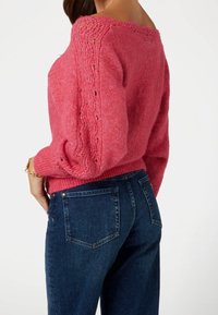 Pull rose avec des détails en dentelle raffinés sur les manches, associé à un jean bleu foncé. Coupe décontractée et ourlet côtelé.