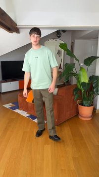 Young man in green t-shirt and brown pants standing on wood floor beside rust-colored couch and large potted plant in modern living room.