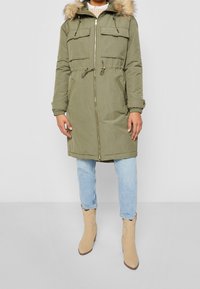 Olivově zelená parka s kapucí lemovanou kožešinou, zipovým zapínáním, stahováním v pase a dvěma předními kapsami. Lehká výplň, tvarované manžety rukávů a střední délka.