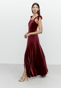 Rochie maxi din catifea bordeaux, cu un corset ajustat, decolteu pătrat și bretele legate. Dispune de o fustă fluidă, cu fante laterale.