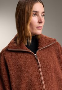 Brown fleece jacket with a high collar, zip closure, and leather accents. Soft texture with a loose design.