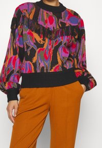 Pull en tricot comprenant une palette de couleurs vibrantes de rose, orange et noir, avec un design à franges sur la poitrine et des poignets côtelés.