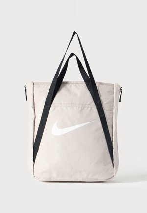 Beige Nike tote tas met groot wit swoosh-logo en lange zwarte schouderbanden, voorvak met rits, rechtopstaand tegen een effen achtergrond.