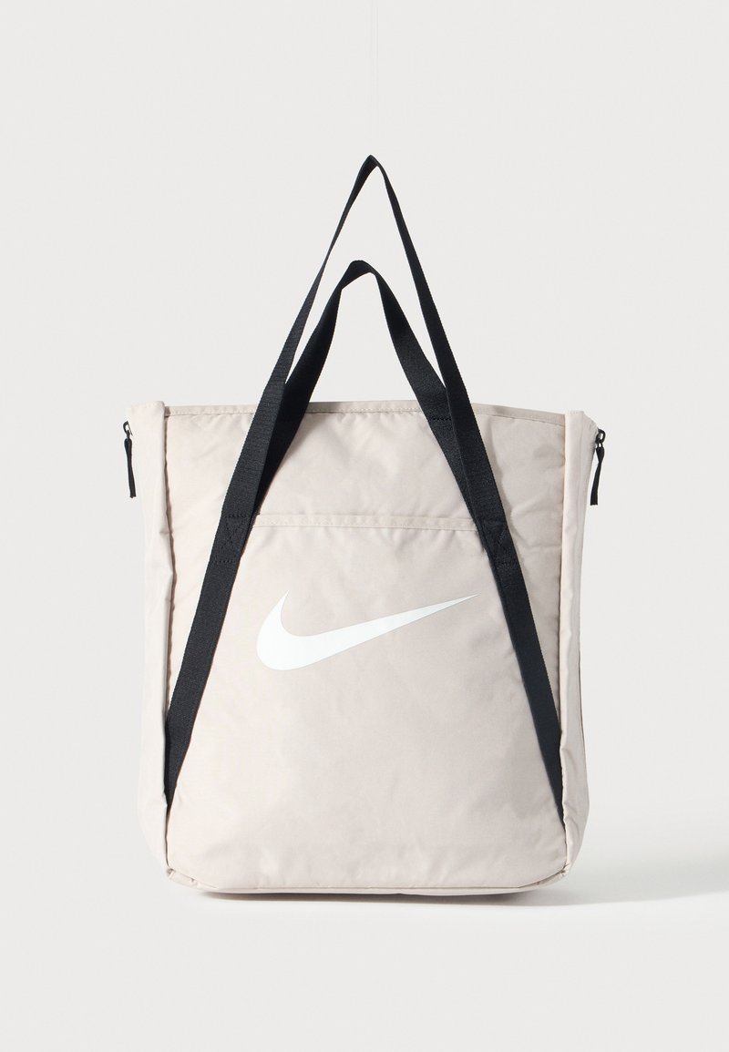 Sac cabas beige Nike avec grand logo swoosh blanc et longues bretelles noires, poche zippée à l'avant, debout sur fond uni.