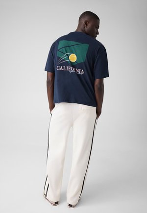 Homme debout, les mains dans les poches, portant un t-shirt bleu marine avec un graphique vert et jaune et un pantalon blanc à rayures noires.
