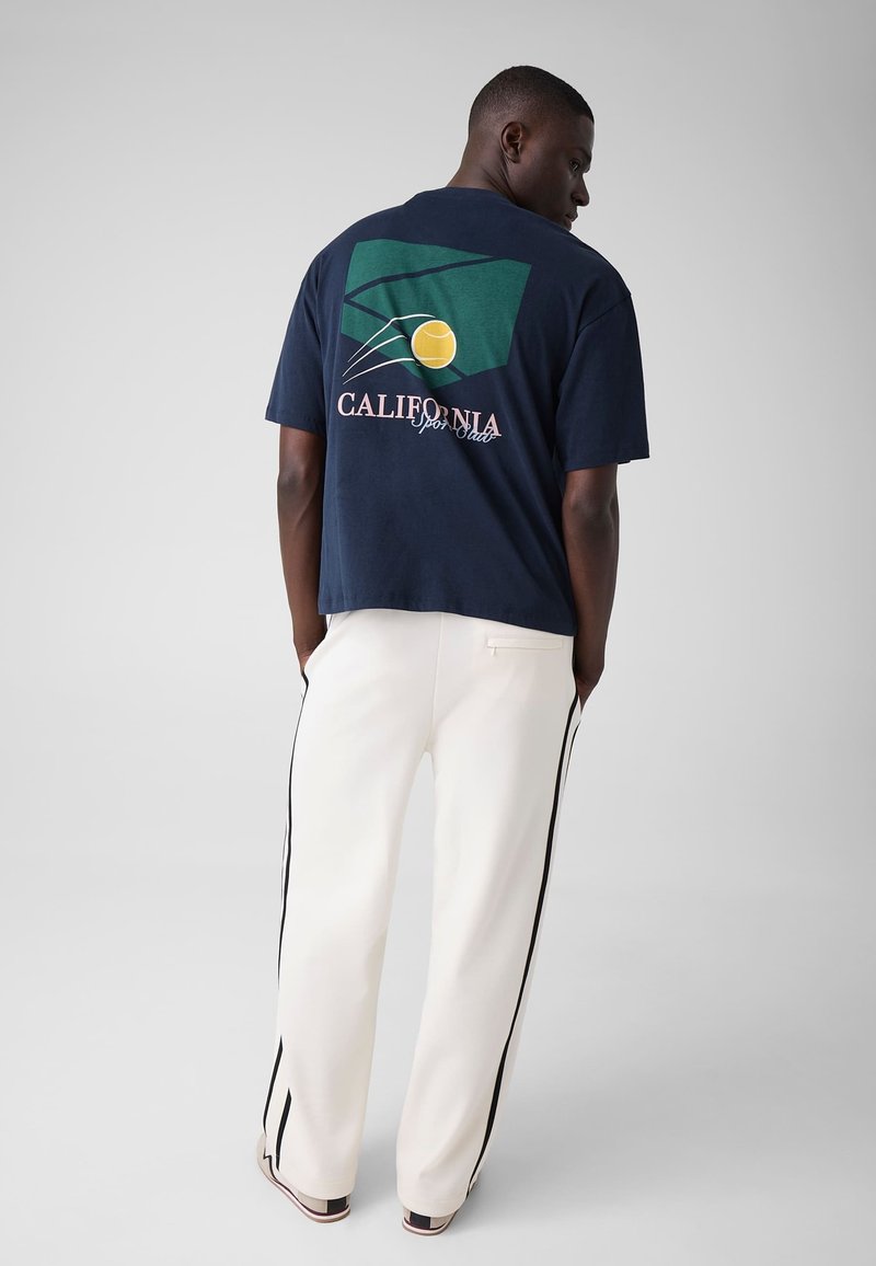 Homme debout, les mains dans les poches, portant un t-shirt bleu marine avec un graphique vert et jaune et un pantalon blanc à rayures noires.