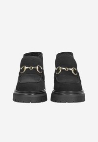 Chaussures en daim noir avec un talon légèrement rehaussé, dotées d'une chaîne en or sur le dessus et d'une semelle en caoutchouc texturée pour une meilleure adhérence.
