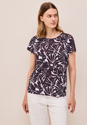 T-shirt à manches courtes en tissu blanc avec des motifs de feuilles violet foncé, col rond et coupe décontractée, associé à un pantalon de couleur claire.