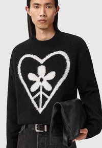 Svart genser med fluffy tekstur, med et stort hvitt hjerte og blomstedesign. Svart belte med skinnt sølvspenne, som holder en svart teksturert veske.