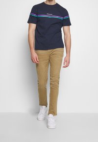 Man i marinblå t-shirt med lila och gröna ränder, beige slim-fit byxor och vita sneakers, som går mot en vit bakgrund.