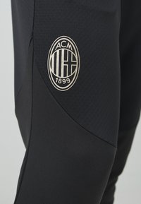 Black athletic pants featuring a textured fabric with a smooth finish, displaying a gold embroidered logo with the text "ACM 1899."