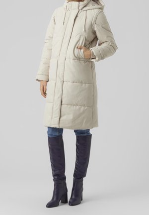 Personne portant un manteau matelassé beige mi-longueur avec capuche, un jean bleu et des bottes noires à talons montant jusqu’aux genoux, debout, une main dans la poche.