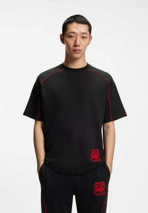 Jeune homme portant une chemise noire à manches courtes et un pantalon avec des coutures rouges et un logo rouge "PR HUGO", debout les mains dans les poches.