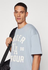 Camiseta de algodón azul claro con mangas cortas, que presenta el texto impreso en blanco "UNDER ARMOUR 96" en una fuente en negrita. El modelo sostiene una bolsa negra.