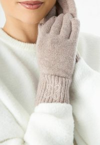 Vrouw in lichtbeige gebreide handschoenen met een geborduurde vlinder op de manchetten, gekleed in een witte donzige trui, die één handschoen dichtbij haar gezicht houdt.