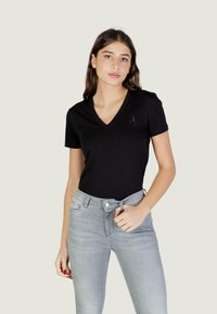 Młoda kobieta w czarnej koszulce z dekoltem w serek z małym detalem w postaci litery "A" oraz jasnoszarych, wysokich dżinsach, stojąca na jednolitym tle.