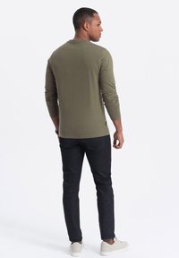 Olivgrünes Langarm-T-Shirt aus weichem Stoff, kombiniert mit dunklen Slim-Fit-Jeans und hellgrauen Sneakers. Sichtbare Nahtdetails am Saum.
