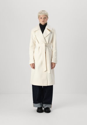 Abrigo de lana blanca con solapas anchas, cintura ceñida con cinturón y bolsillos laterales, combinado con jeans de pierna ancha oscuros y zapatos negros de tipo slip-on.