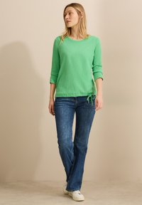 Camicia a manica lunga verde chiaro con texture, scollo rotondo e dettaglio con nodo laterale, abbinata a jeans blu e sneakers bianche.