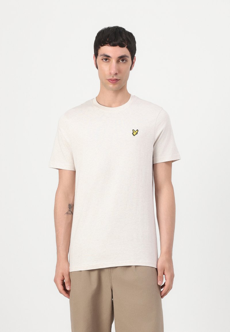 T-shirt en coton beige clair à manches courtes et col rond. Présente un petit logo de papillon jaune sur la zone gauche de la poitrine.
